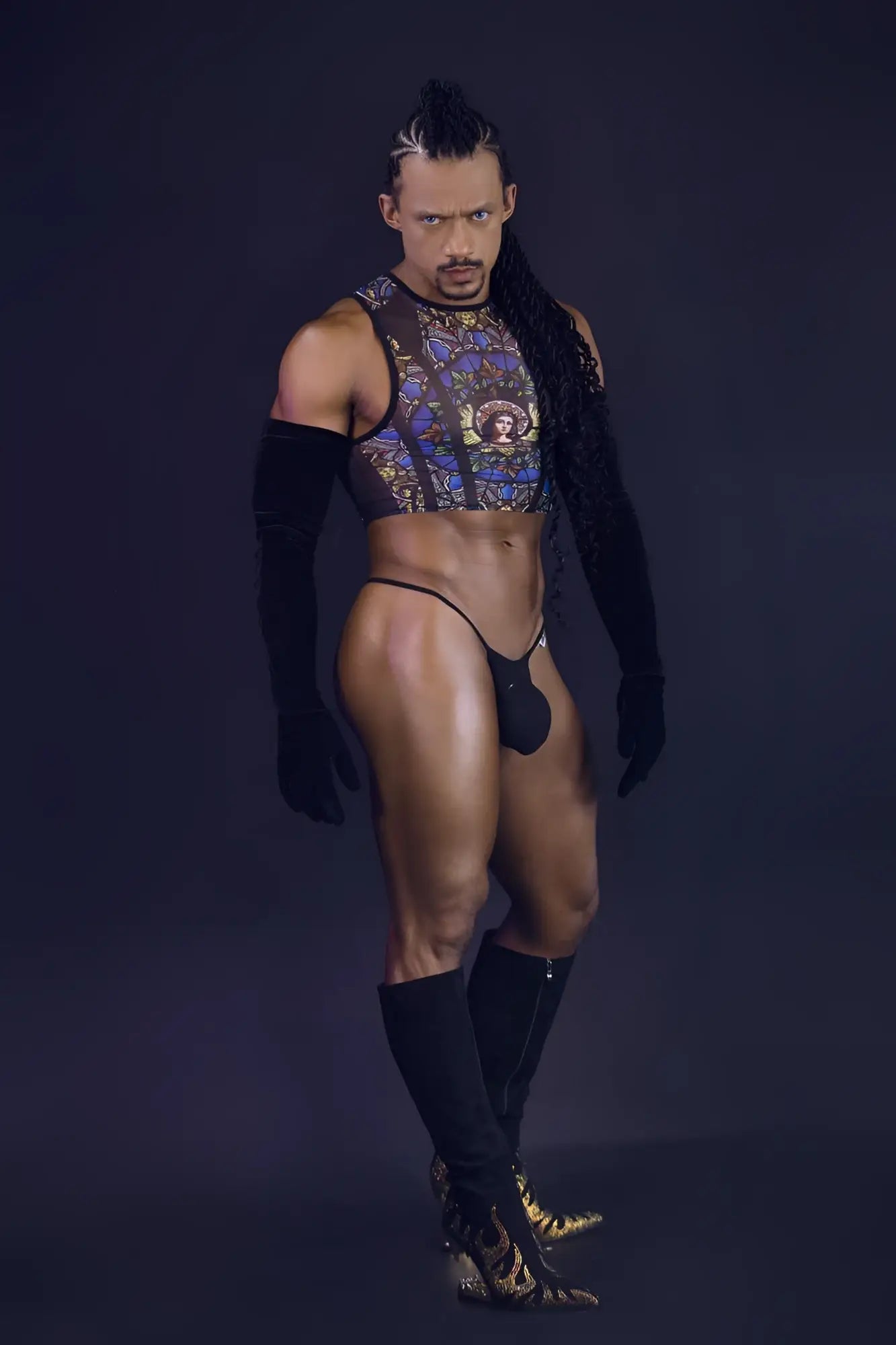 A man wearing a sheer stained glass patterned black crop top with gold and blue designs over black arm sleeves