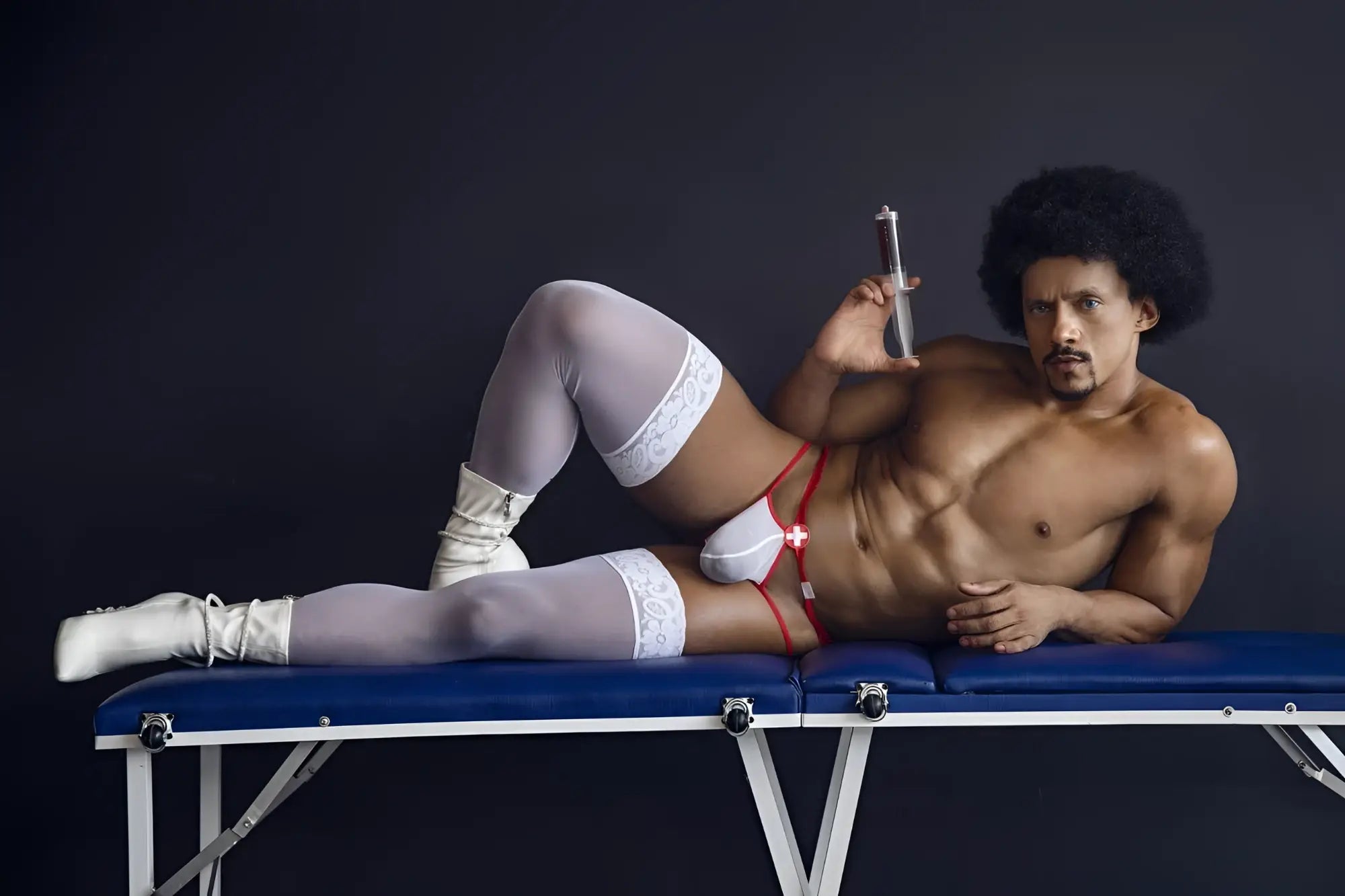 A muscular man with an afro lies on a blue table wearing candyMan 99833 murse thongs red-white with white lace stockings