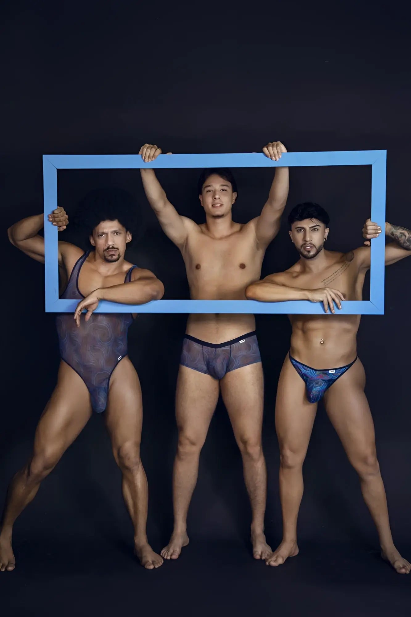 Three men holding a blue rectangular frame featuring CandyMan 99873 seawater trunks with multicolor print