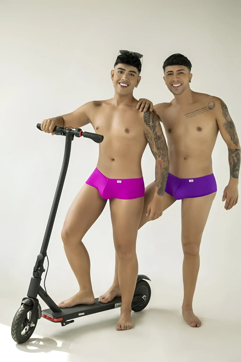 Two men wearing orchid-colored trunks pose together, one standing on an electric scooter against a white background.
