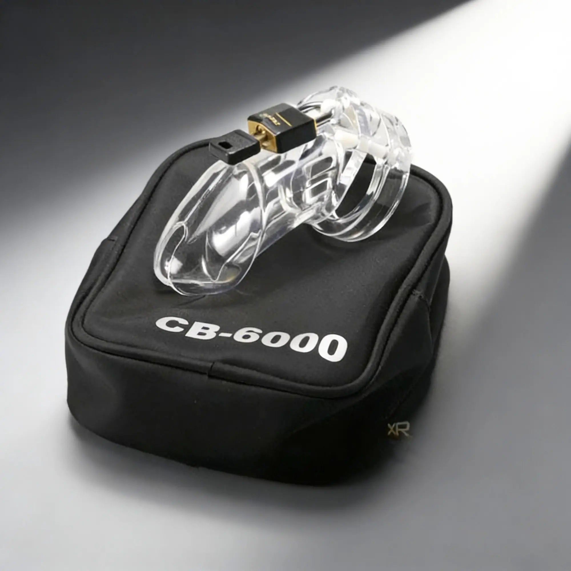 Clear plastic CB-6000 male chastity device with a small black padlock, resting on a black labeled storage pouch.