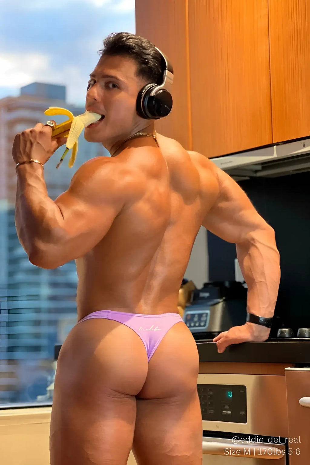 A muscular man wearing black and silver over ear headphones eating a banana featuring cheeky classic g string thong digital violet