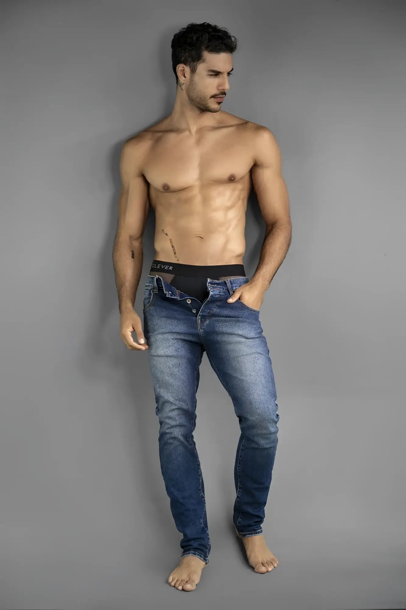 A shirtless man wearing Clever 26059 Tanatos black briefs with a branded waistband under unbuttoned blue jeans.