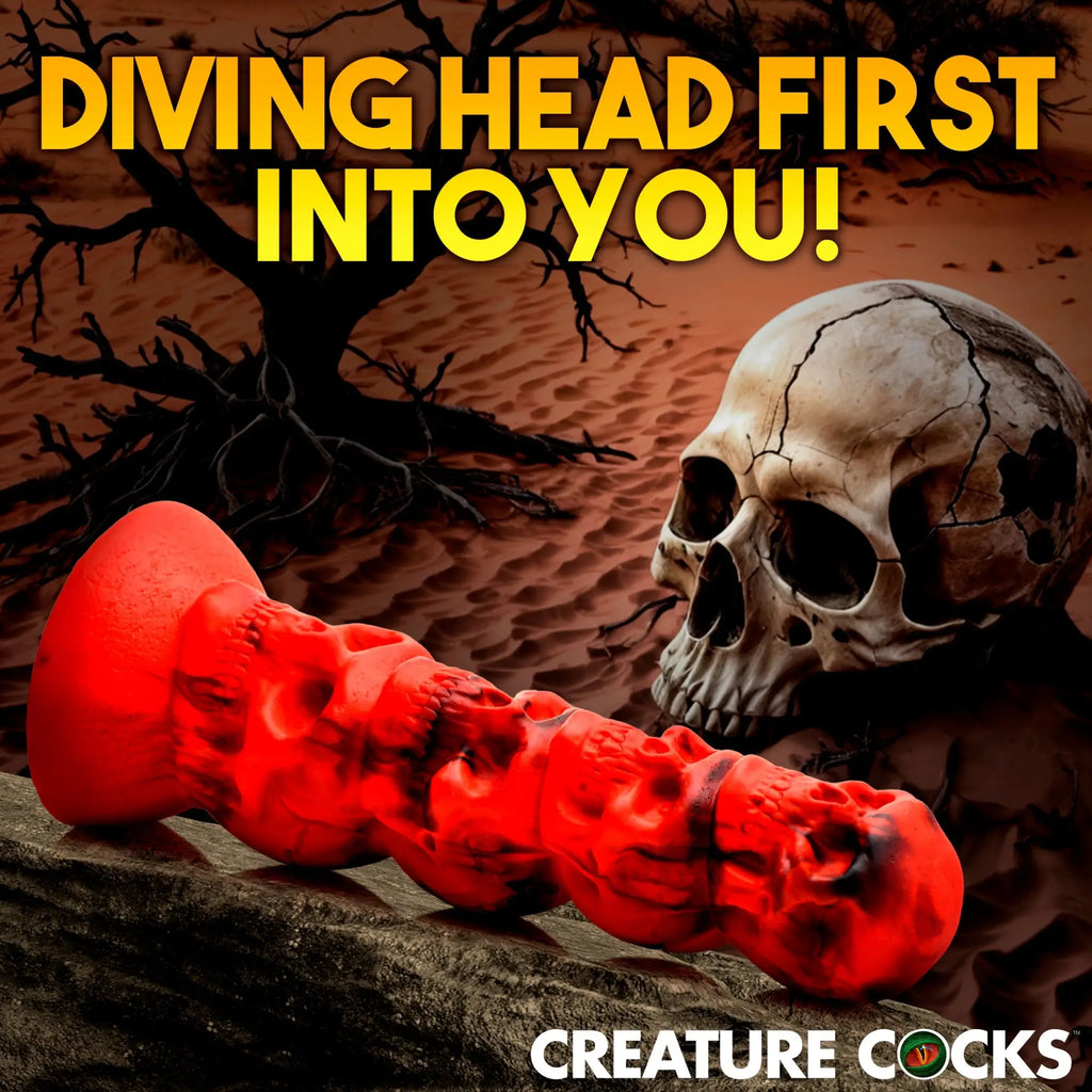 Creature Cocks Doom dildo in red and black, shaped like a stack of skulls, placed before a human skull in a desert.