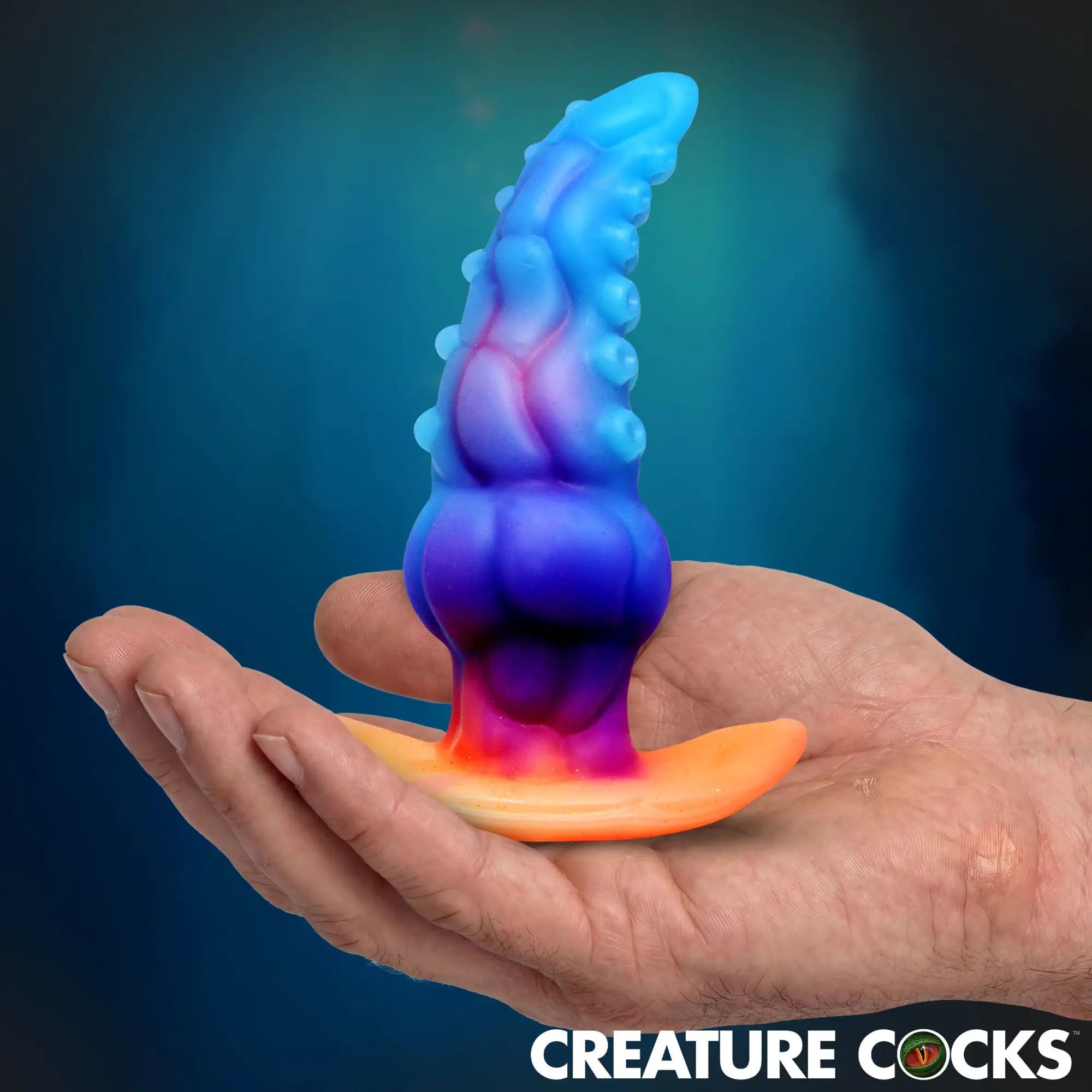 Creature Cocks Glowtopus silicone anal plug with tentacle texture, shifting from blue to orange in a palm-up hand.