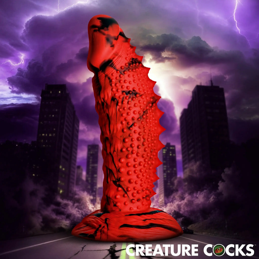 Creature Cocks Gojira red and black marbled silicone dildo with textured spikes, standing against a stormy city background.