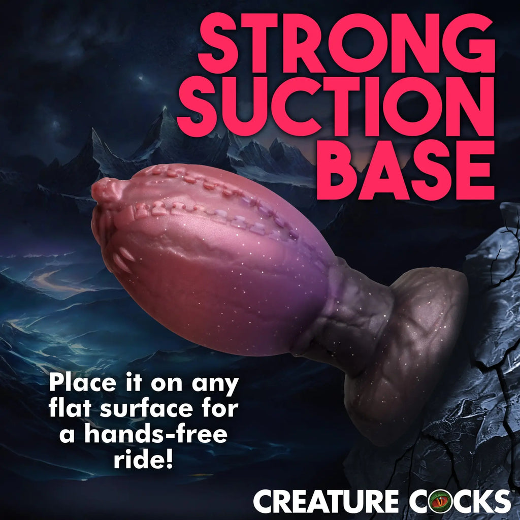 Creature Cocks Large Dragon Hatch silicone egg in purple with a black suction base against a fantasy mountain backdrop.