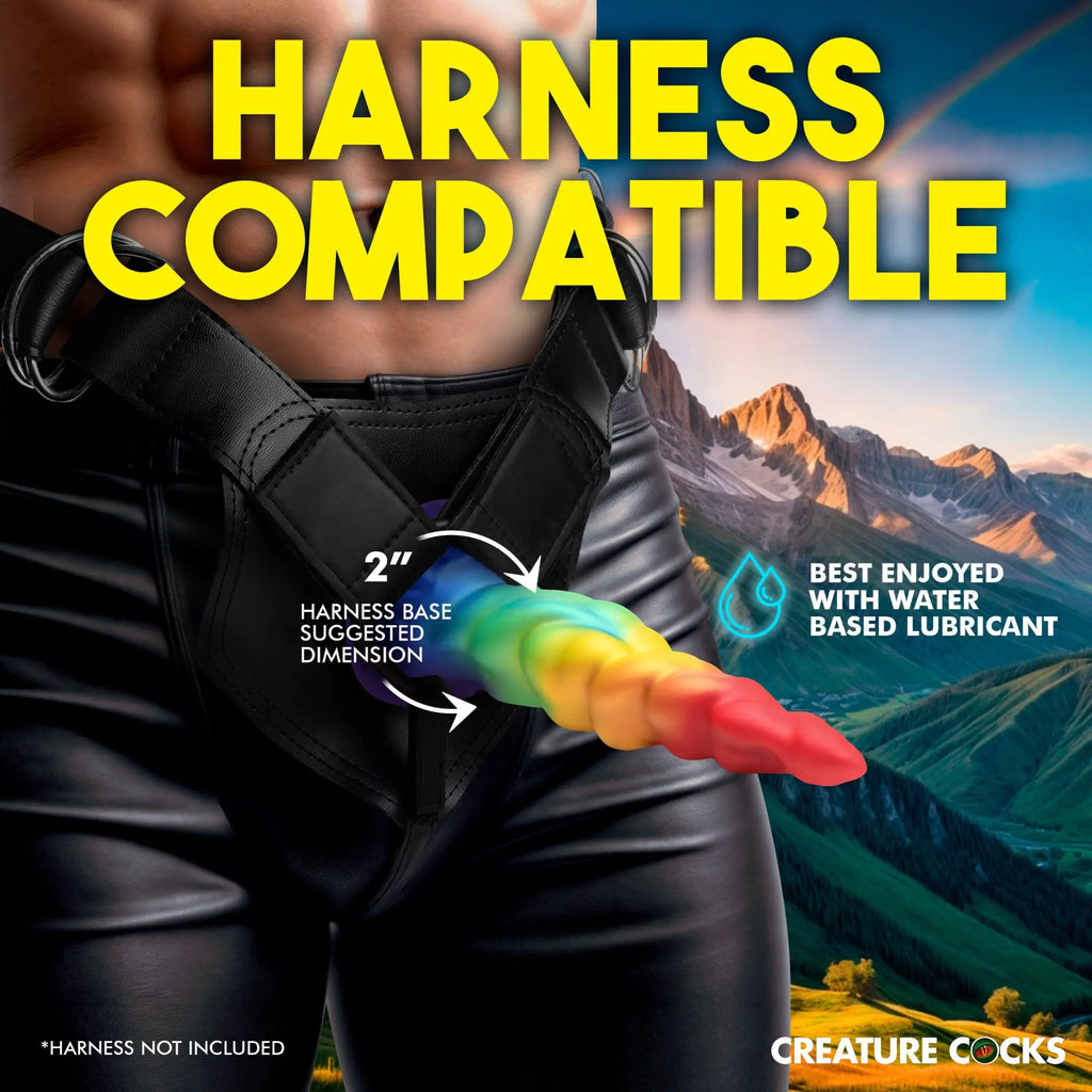 Creature Cocks Magic Rider rainbow silicone unicorn dildo in a black leather harness against a mountain background.