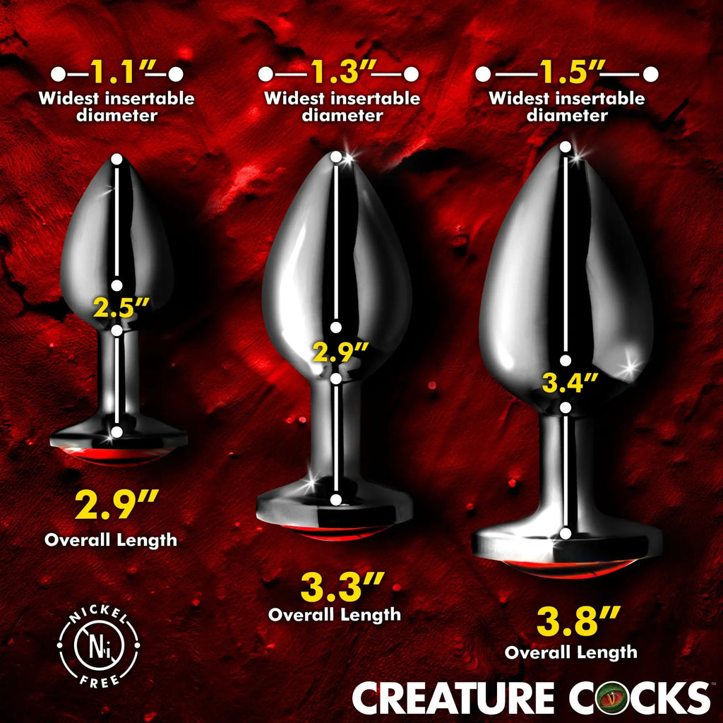 Three sizes of silver metal Creature Cocks Sinister Eye butt plugs with dimension labels on a dark red background.