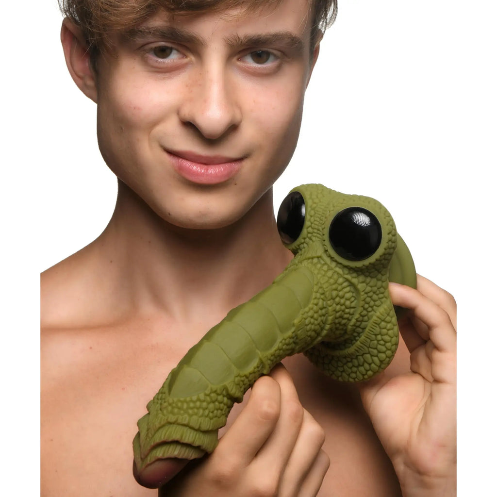 A man holds a green, scaly Creature Cocks Swamp Monster silicone dildo featuring large, black, bug-like eyes.
