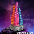 Creature Cocks Tenta-Trio dildo: A triple-tentacle silicone toy in orange, pink-purple, and teal against a sea backdrop.