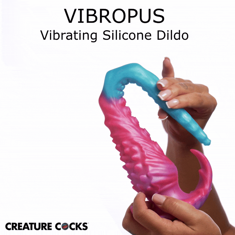 A hand holds the Creature Cocks Vibropus, a curved, textured blue and pink silicone vibrating dildo on a white background.