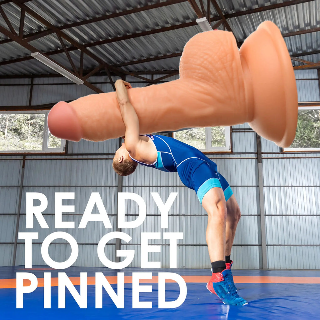 A wrestler lifts a giant, skin-toned Curve Toys Wrestling Wayne 7-inch dildo with a suction cup base on a blue mat.