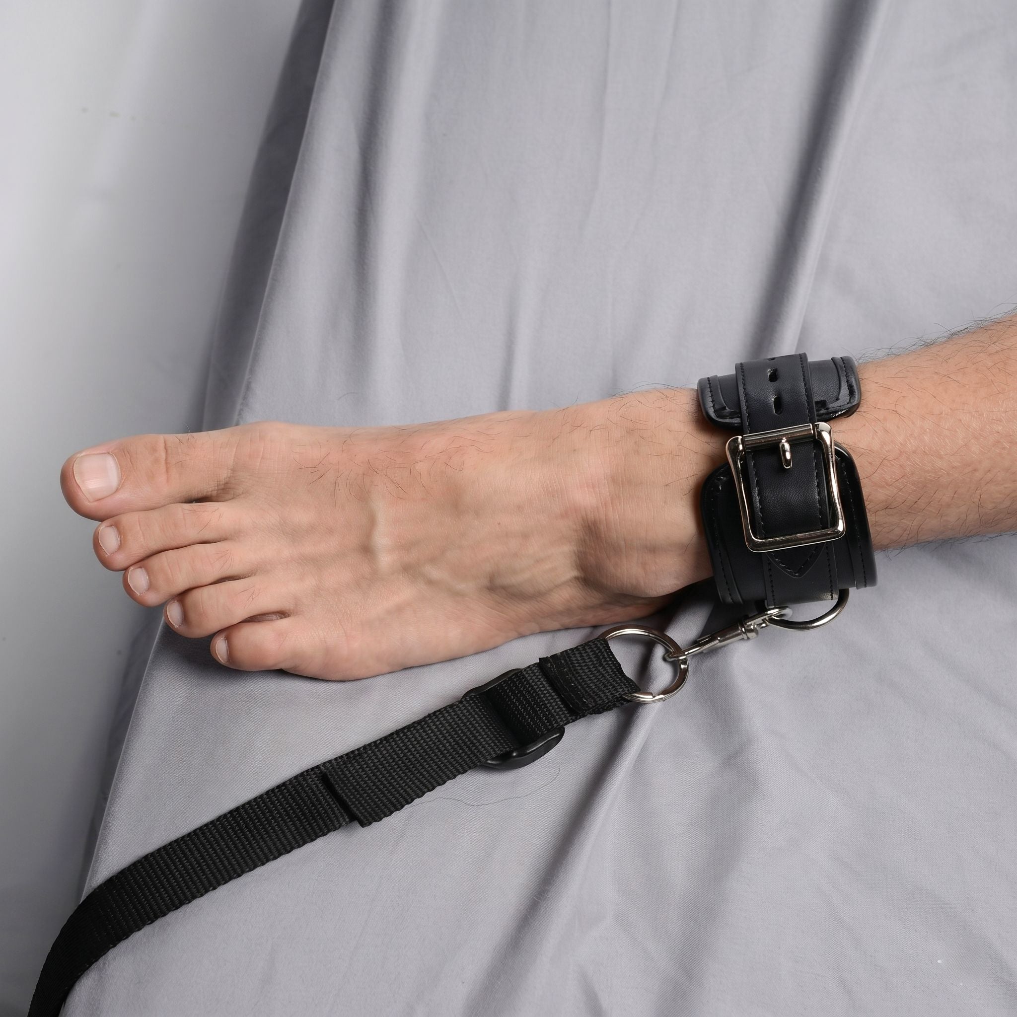 Strict Deluxe Bed Restraint System: Black leather ankle cuff with a silver metal buckle and an attached black strap.