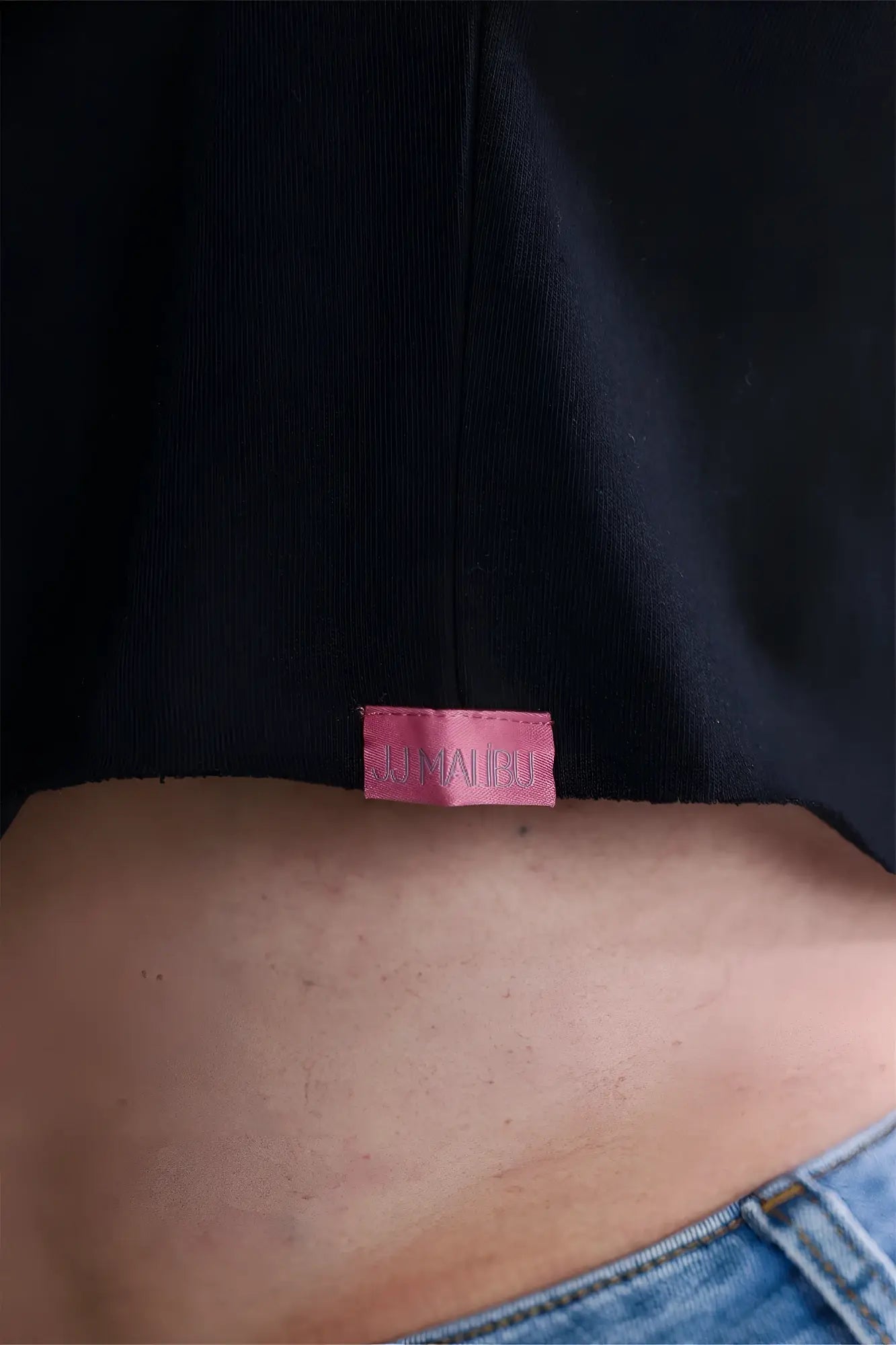 Pink rectangular kumaipoo label with stitched edges sewn on the back of a black escapism muscle-fit crop tank puta