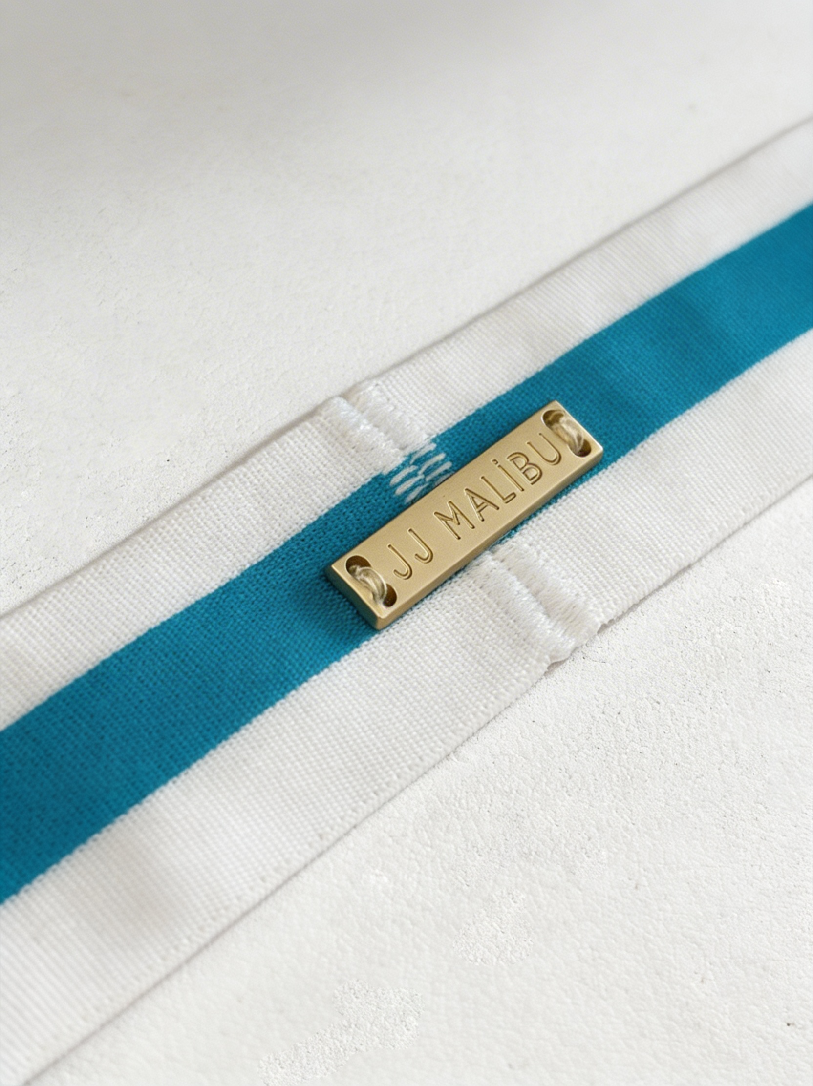 Blue on-display jockstrap with white fabric strap featuring bold teal stripes and gold-tone metal tag engraved jj malibu