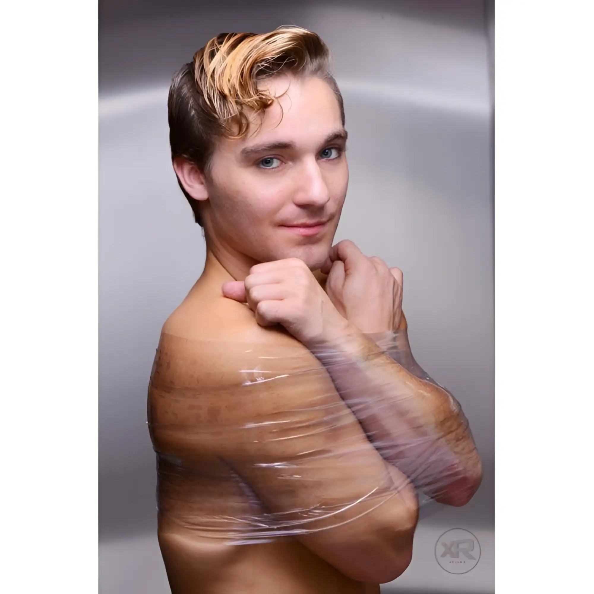 A man with blond hair poses with Frisky Clear Bondage Tape wrapped securely around his bare upper body and arms.