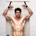 Muscular man wearing black Frisky Over the Door Entryway Restraints, arms raised against a white door.