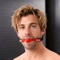 A man wears the Frisky Silicone Comfort Ball Gag, featuring a bright red silicone sphere on a black adjustable strap.