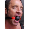 Frisky Subdue Me Breathable Ball Gag with a black perforated ball on a red and black leather strap worn by a man.