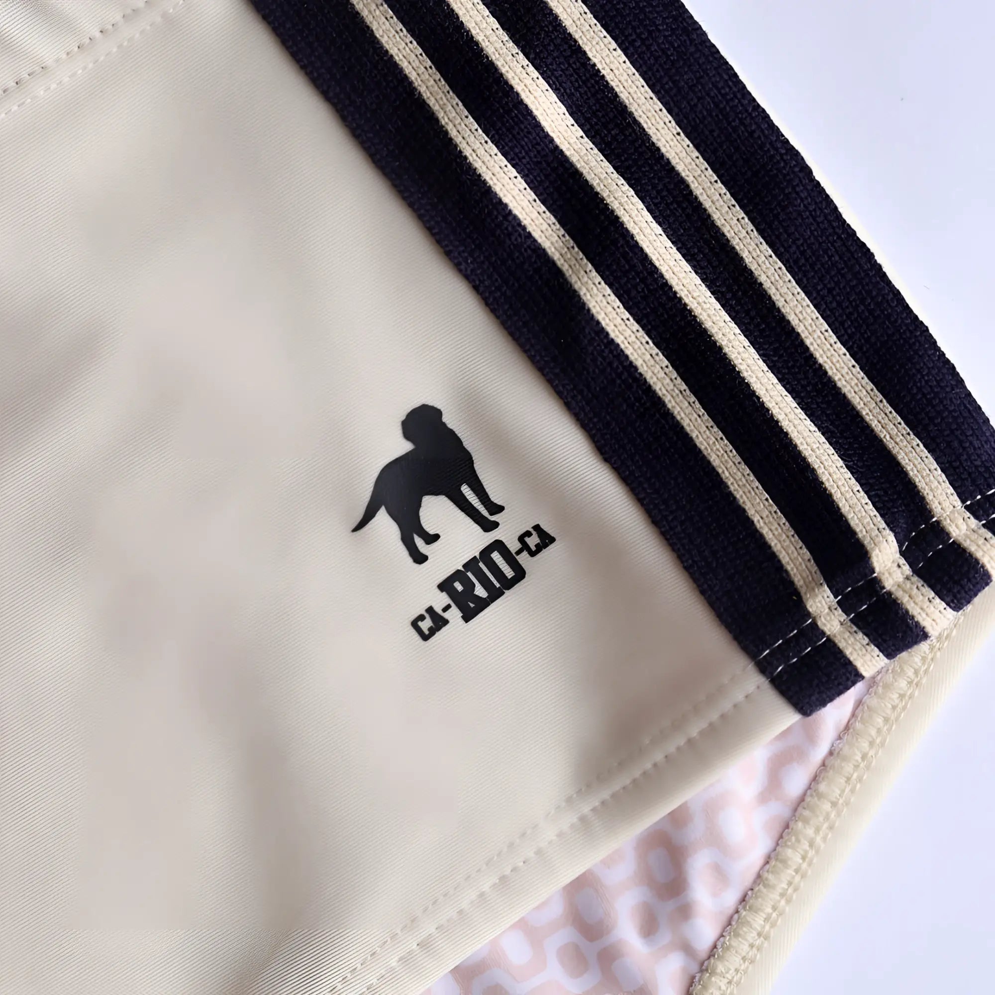 Cream-colored galão creme sunga athletic shorts with black and beige striped side panels and embroidered dog logo