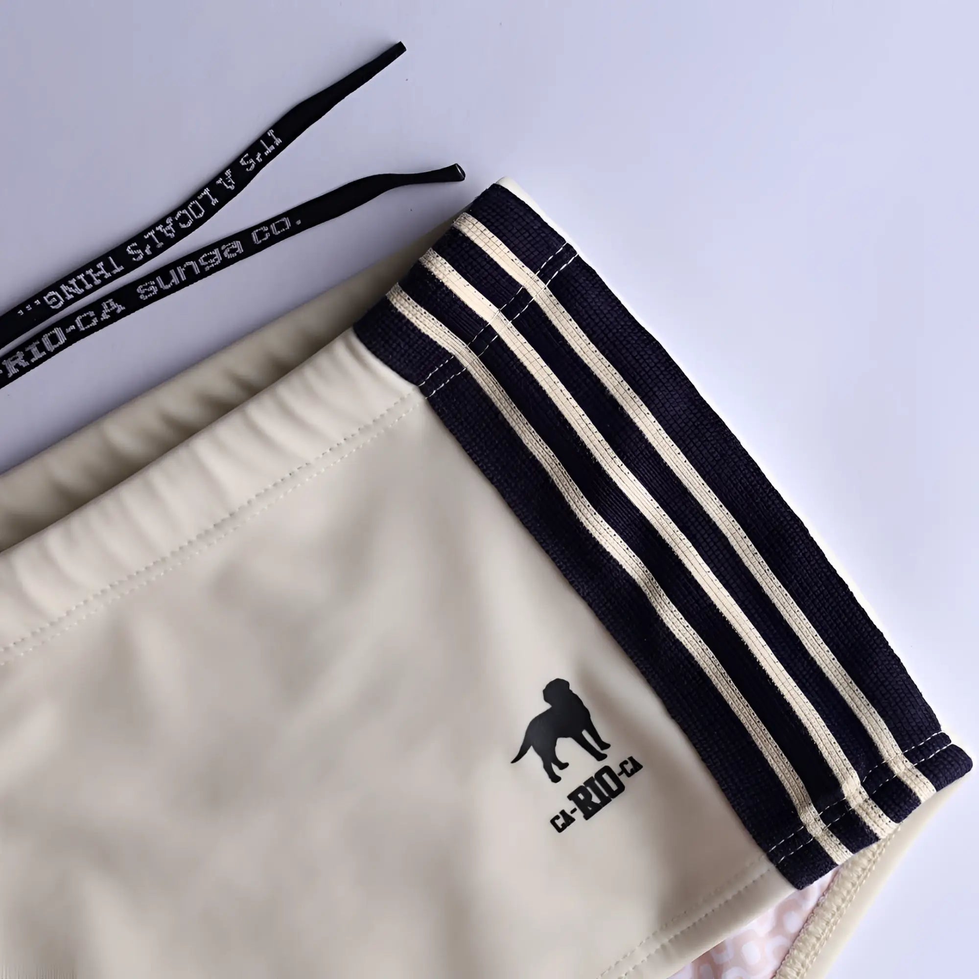 Cream-colored galão creme sunga designer swim shorts with black side stripes and small black dog logo on the leg