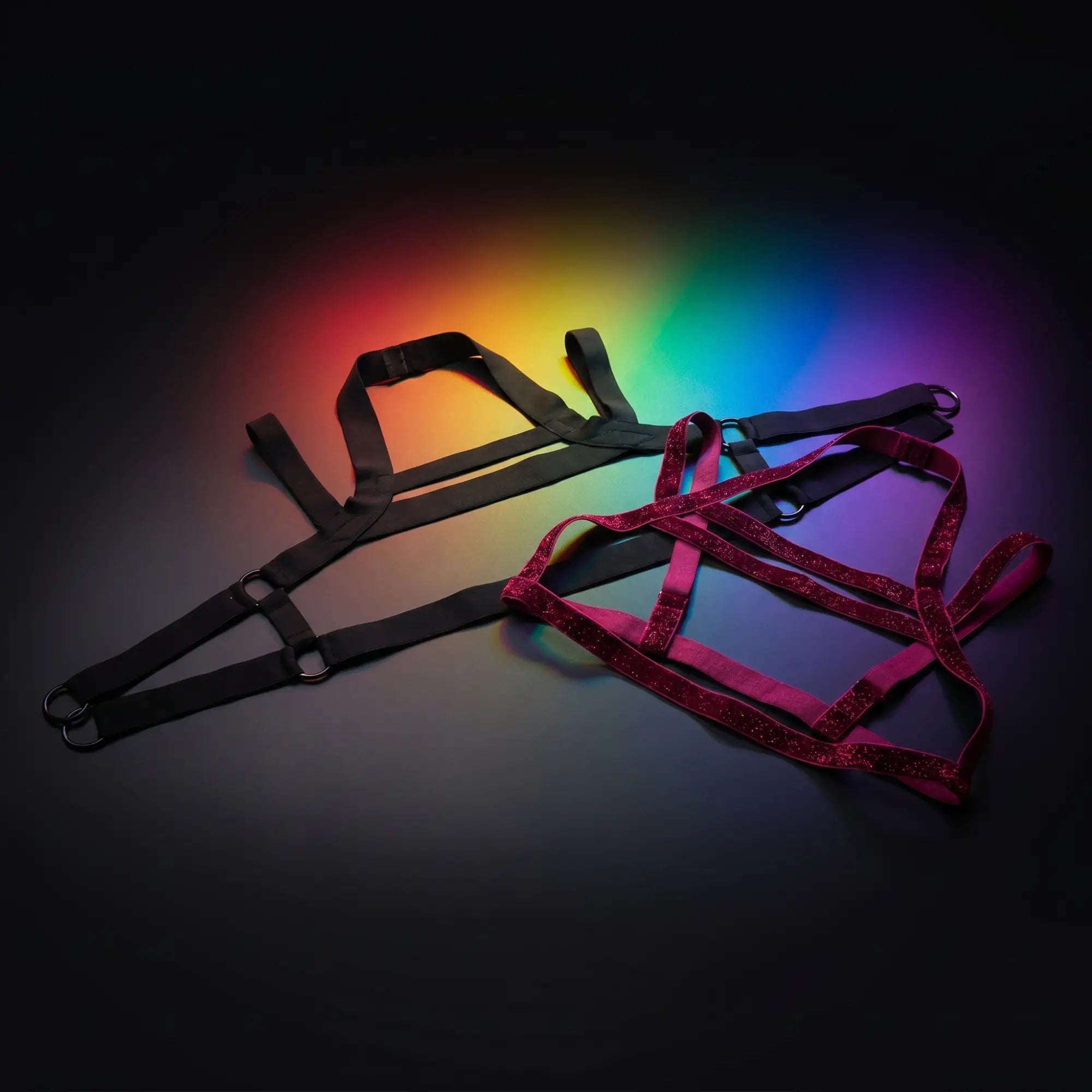 Black multi-band cage harness and raspberry glitter harness flat lay on dark surface with rainbow studio lighting