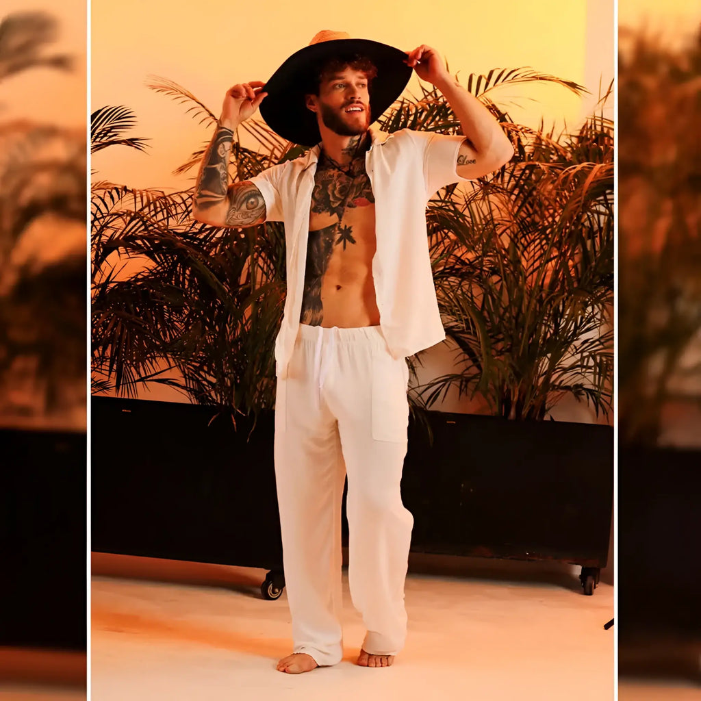 A man wearing gigo chalis beige loose-fitting pants paired with a wide-brimmed black hat and an open white shirt revealing tattoos