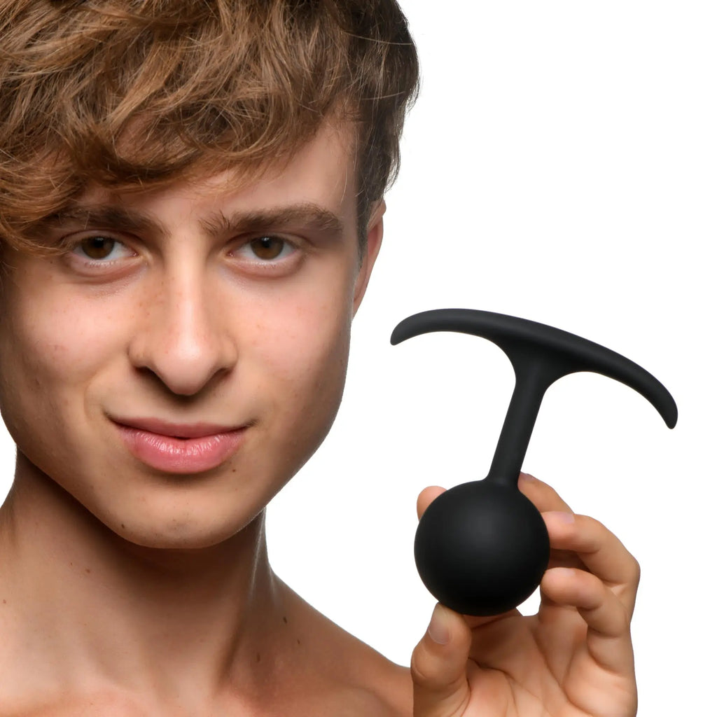 A person holds a smooth, black Heavy Hitters Premium Silicone Weighted Anal Plug with a T-shaped handle.