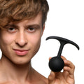A person holds a smooth, black Heavy Hitters Premium Silicone Weighted Anal Plug with a T-shaped handle.