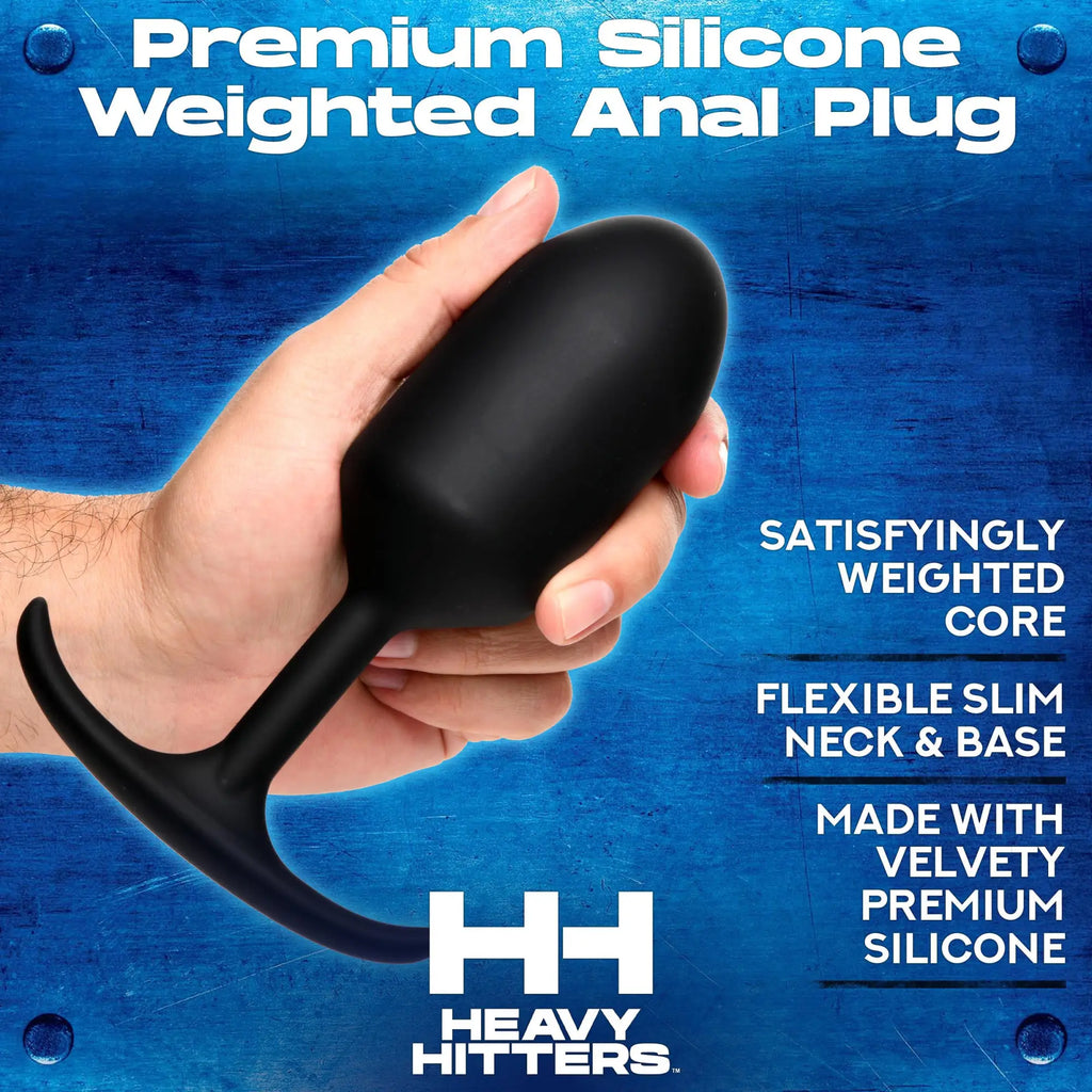 A hand holds a smooth, black Heavy Hitters Premium Silicone Weighted Bullet Anal Plug against a blue metallic background.