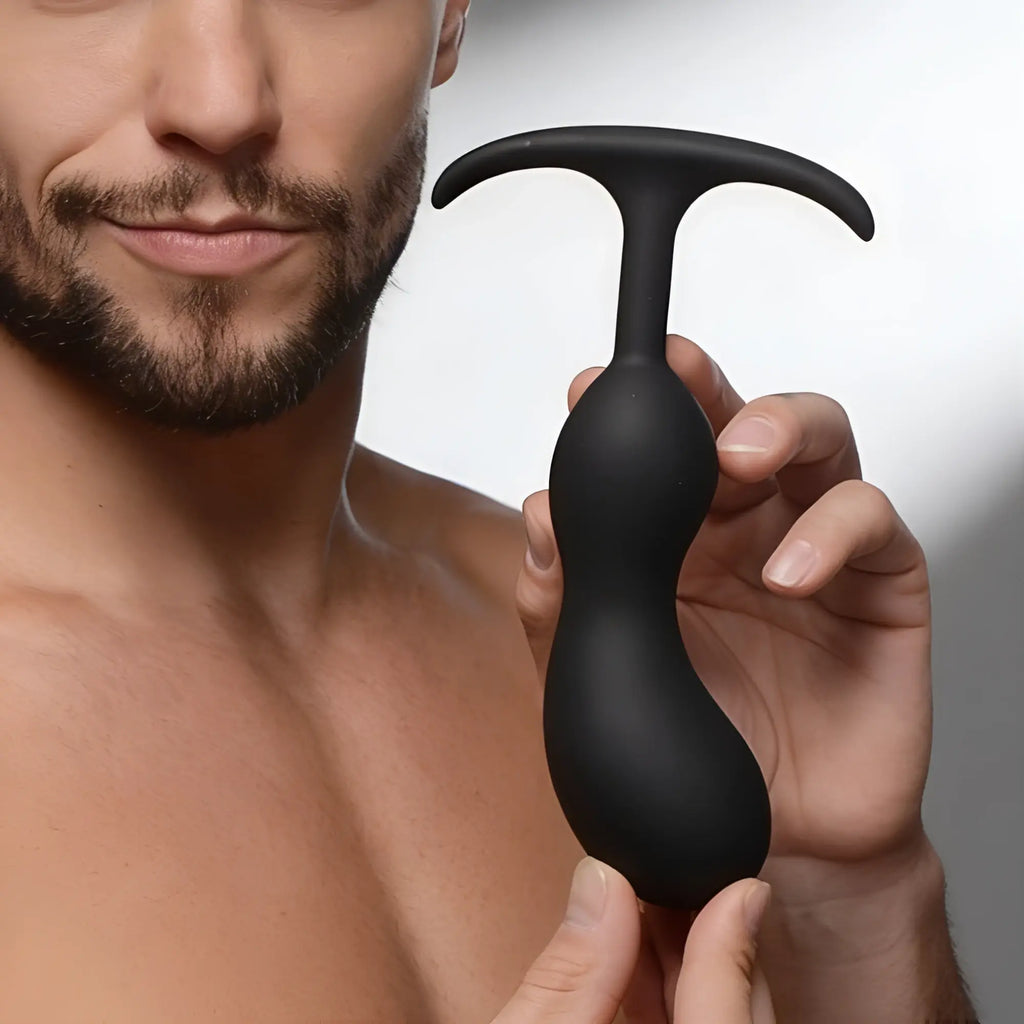 A man holds a smooth, matte black Heavy Hitters silicone prostate plug with a curved base against a light background.