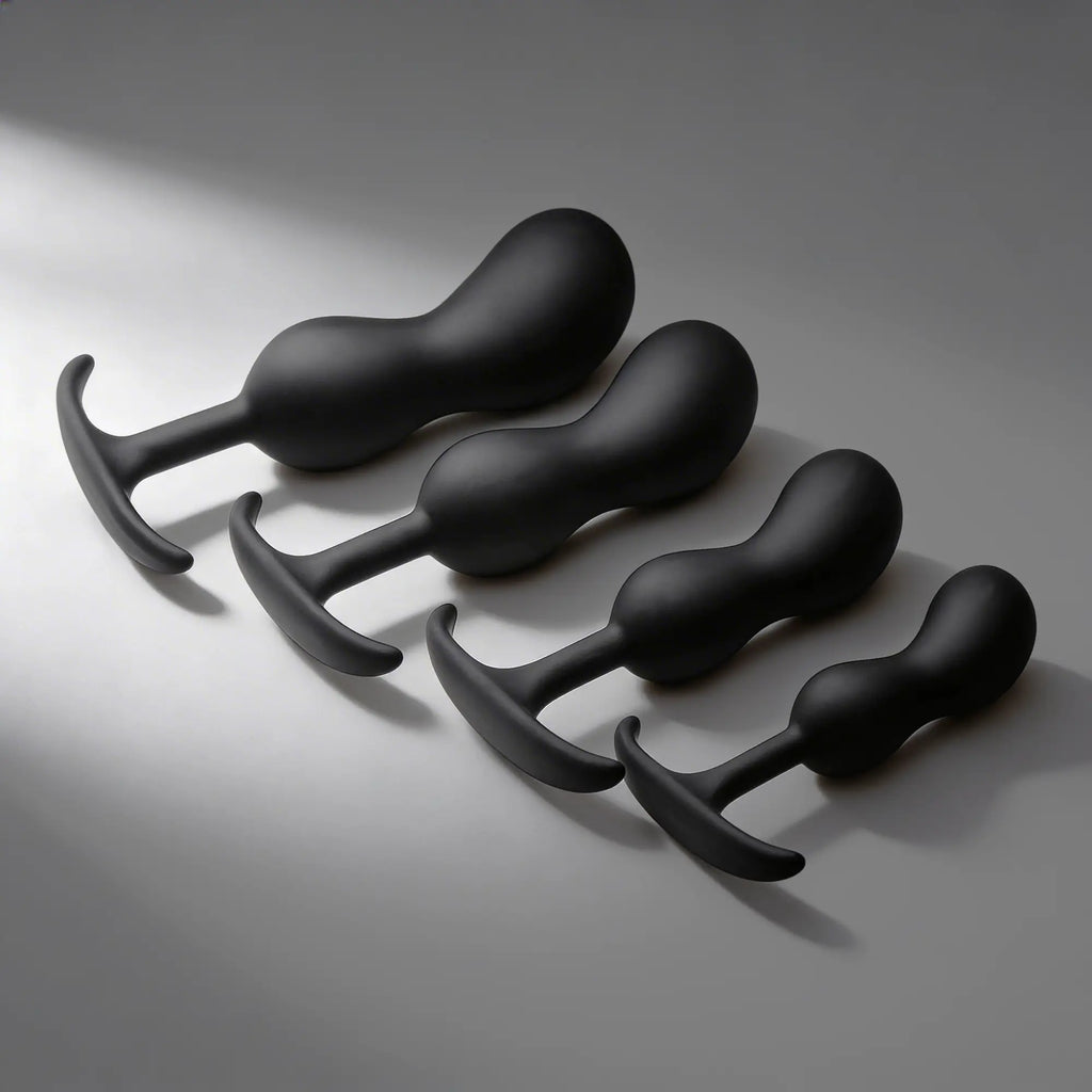 Four matte black Heavy Hitters silicone prostate plugs in graduated sizes, arranged diagonally on a light grey surface.