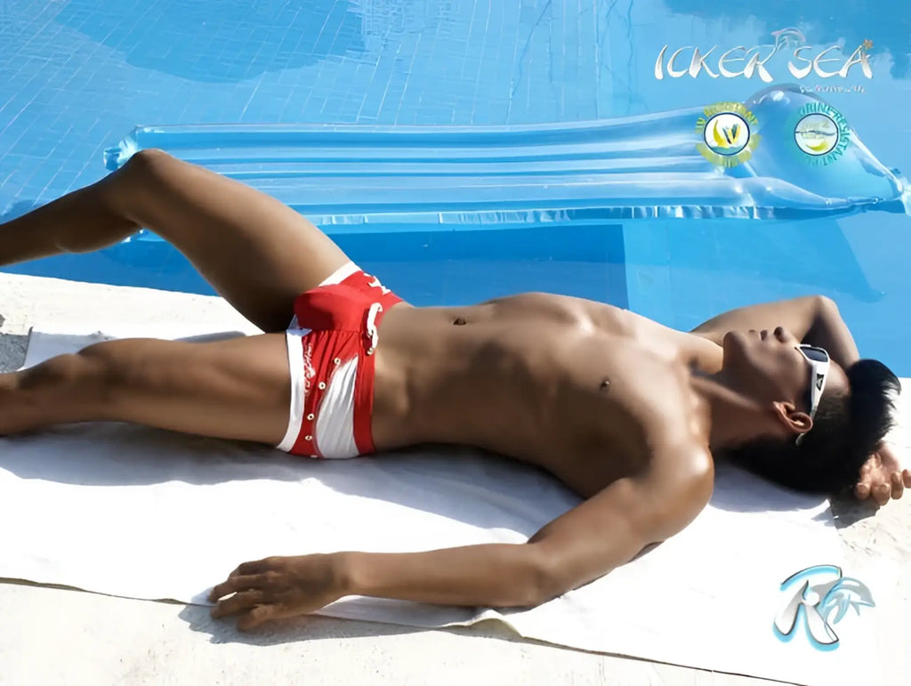 A man lies on a towel by the pool wearing icker sea eyelets square cut red swim trunks with silver buttons