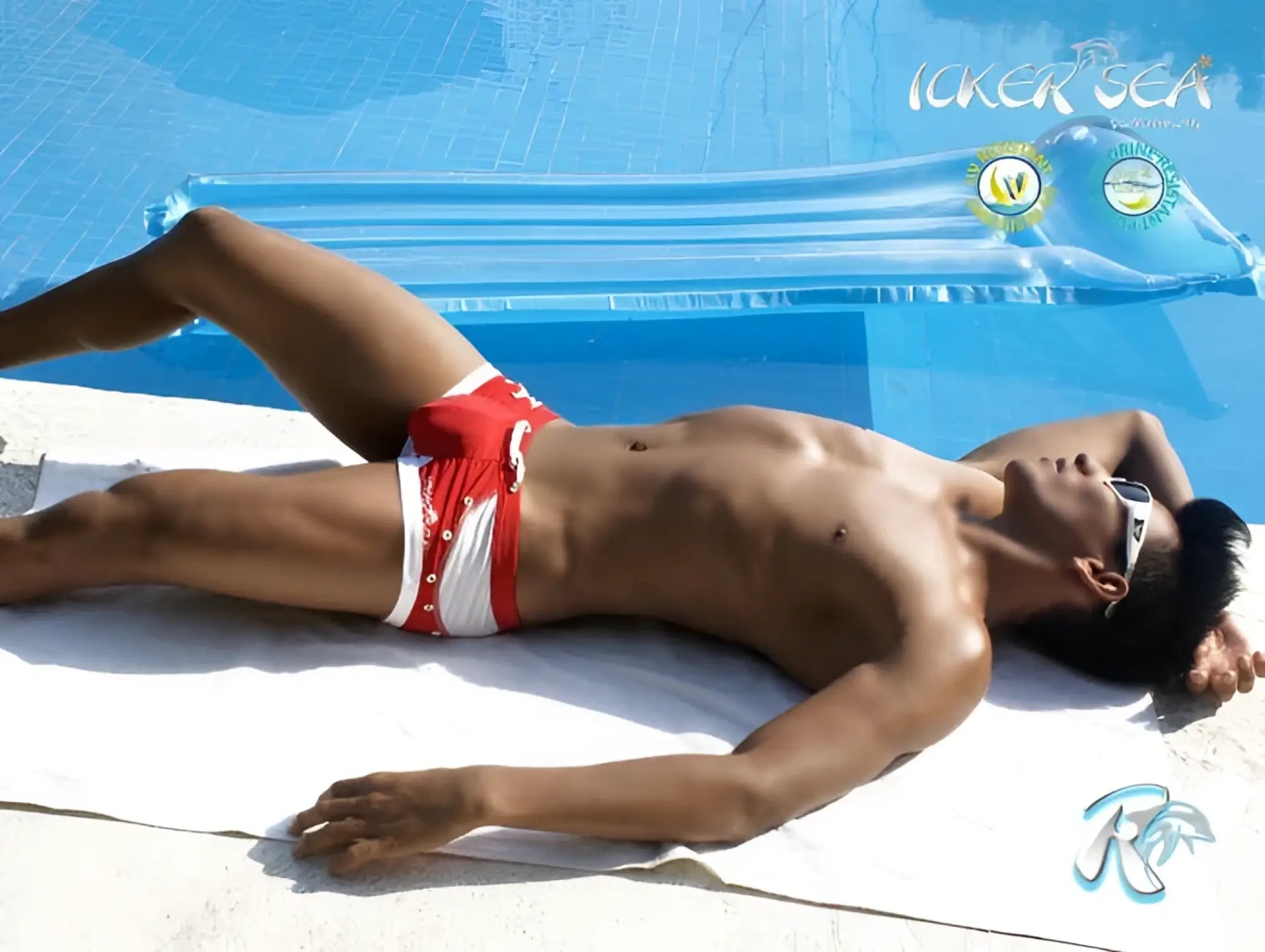 A man lies on a towel by the pool wearing icker sea eyelets square cut red swim trunks with silver buttons