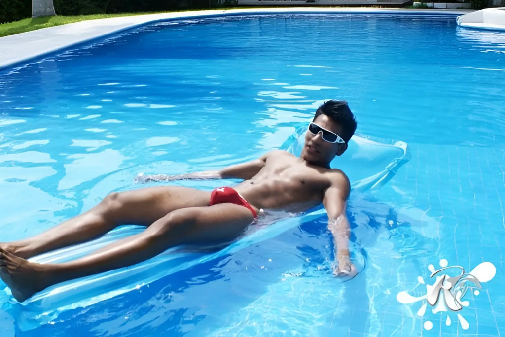 A man wearing icker sea lifeguard micro slip bikini swimwear red floats on a blue inflatable pool float in clear water