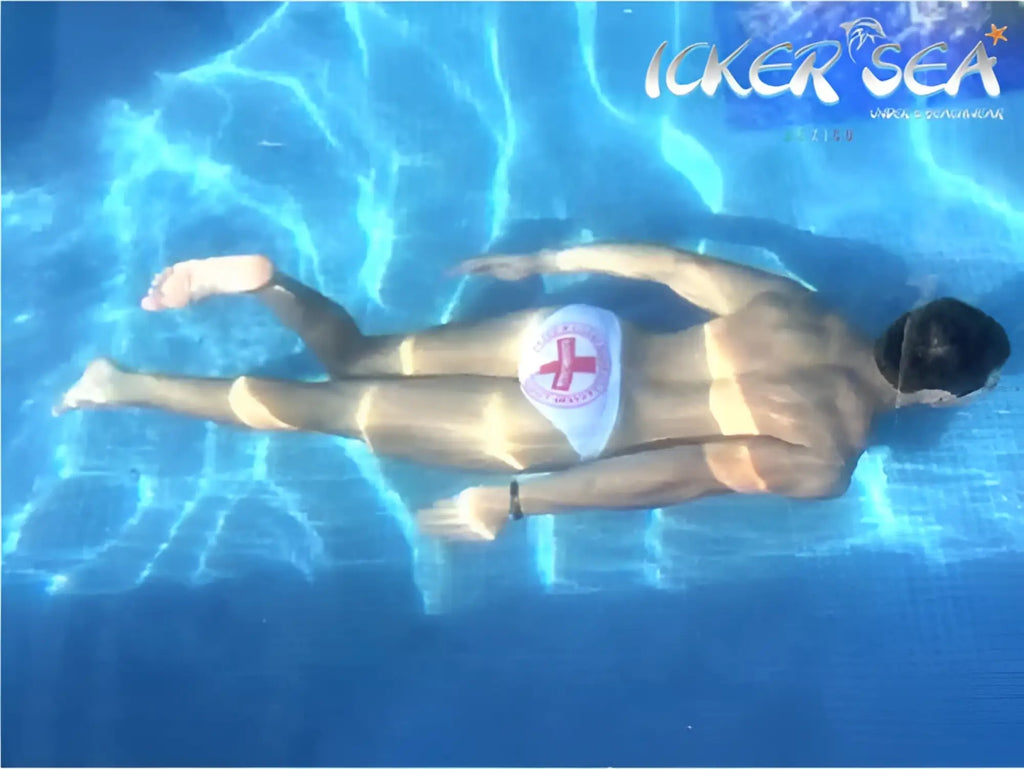 A man floats underwater wearing icker sea lifeguard micro slip bikini swimwear white with red cross emblem