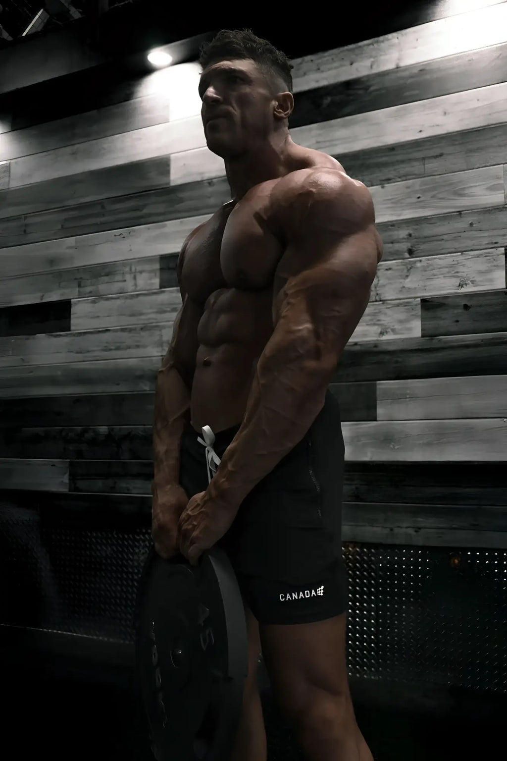 A muscular man wearing black agile bodybuilding 4 inch shorts with zipper pockets and canada logo holding a weight plate