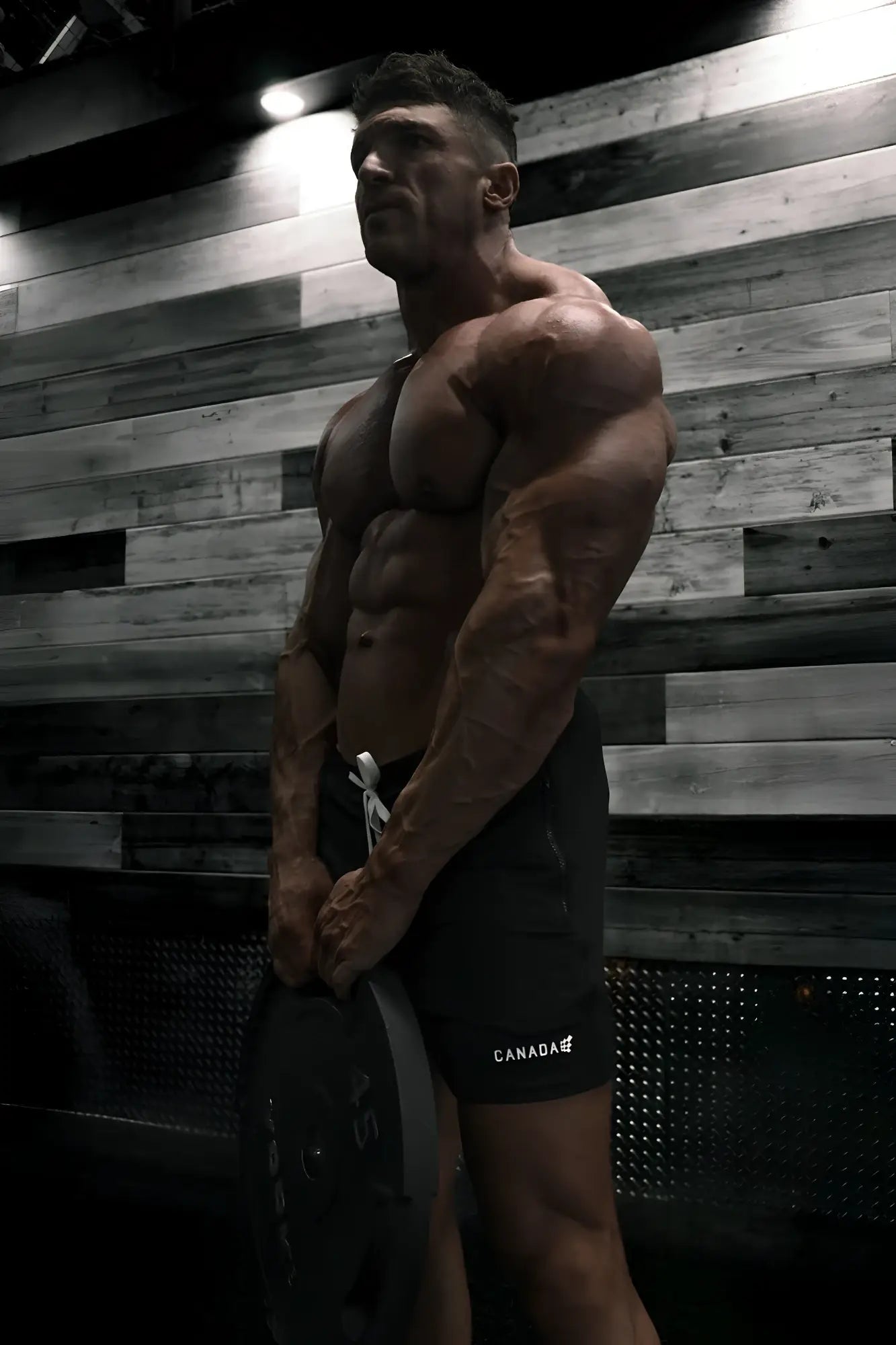 A muscular man wearing black agile bodybuilding 4 inch shorts with zipper pockets and canada logo holding a weight plate