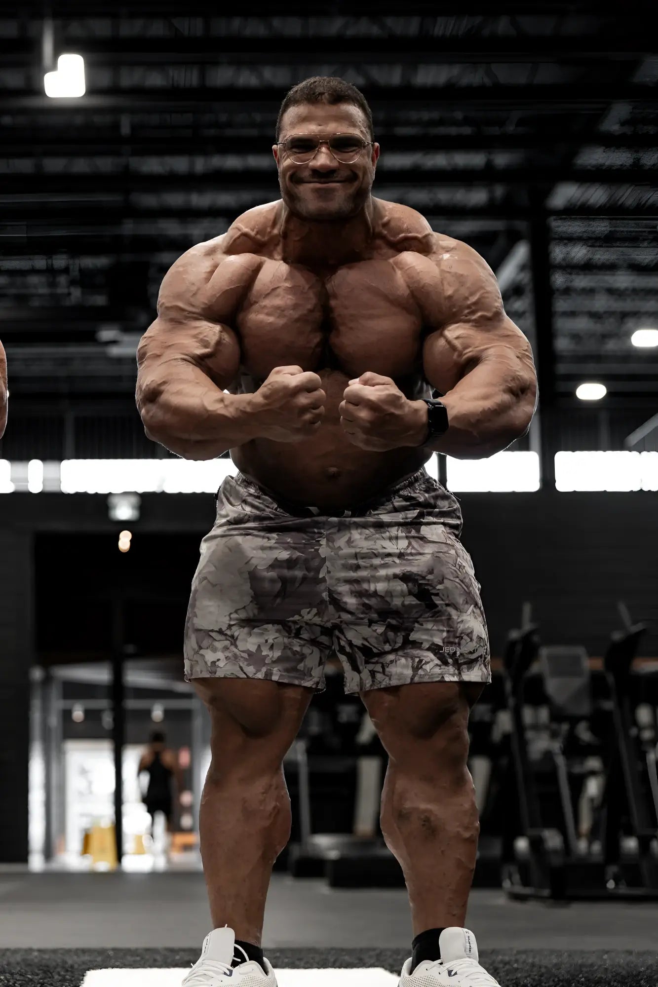 A muscular man wearing agile bodybuilding 4 inch leaf camo shorts with zipper pockets and white sneakers