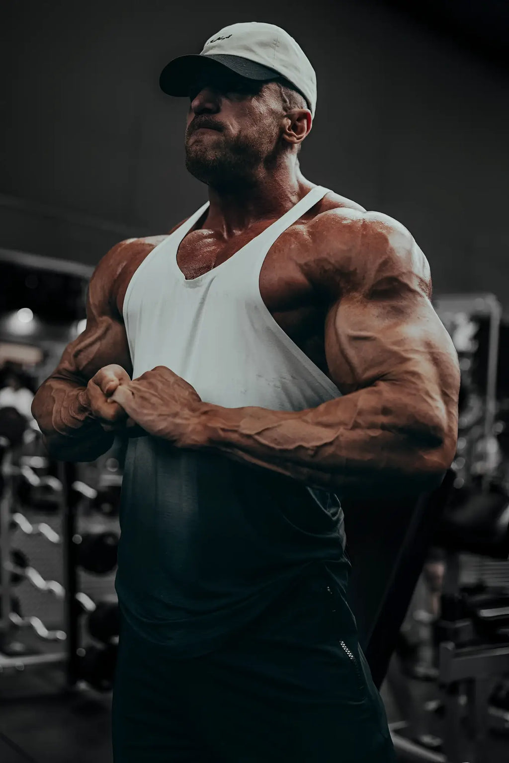 A muscular man wearing a white tank top and light baseball cap modeling a black ombre classic bodybuilding racerback stringer