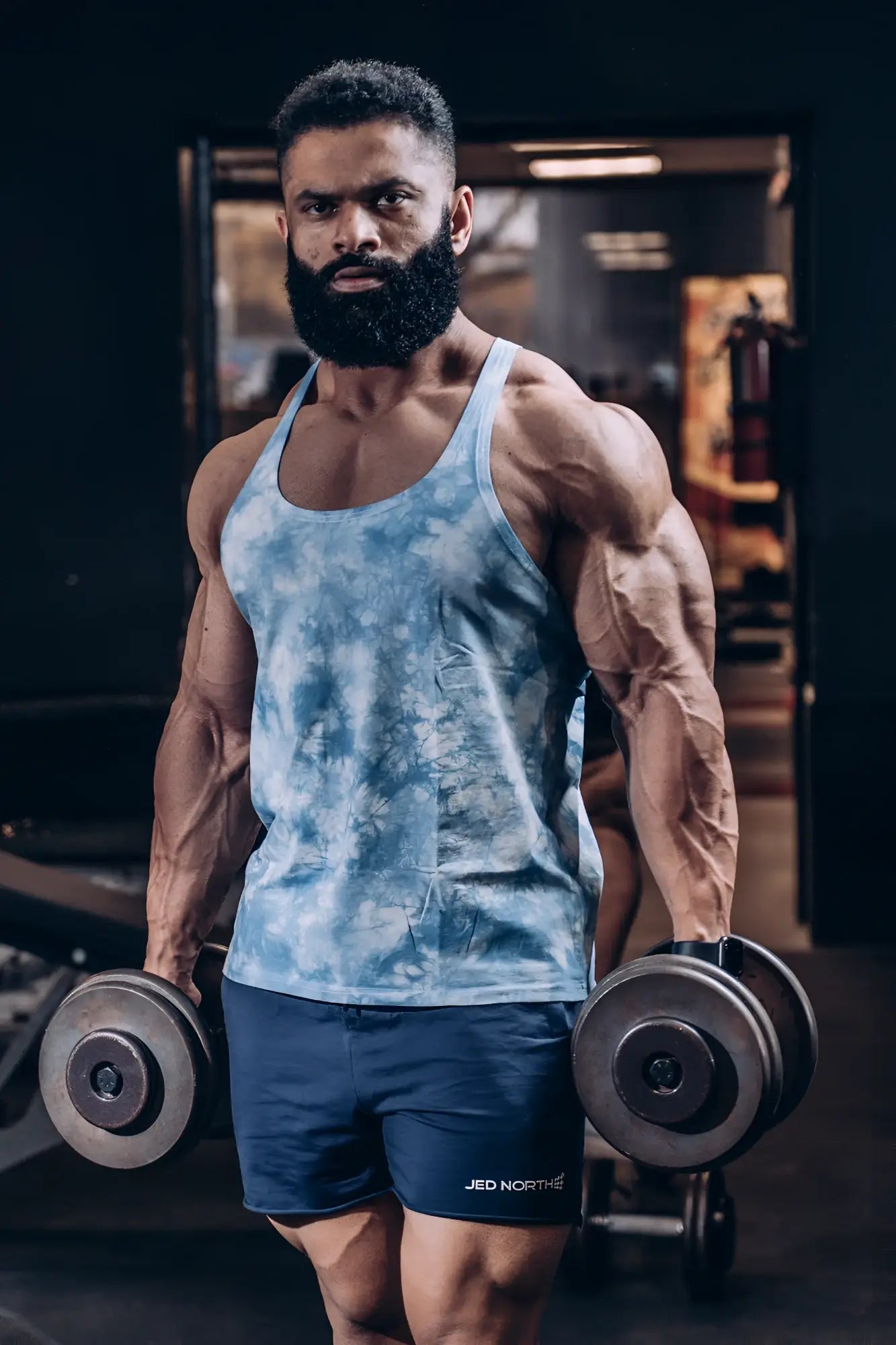A man wearing jed north classic bodybuilding racerback stringer in gray tie dye and navy shorts holding dumbbells