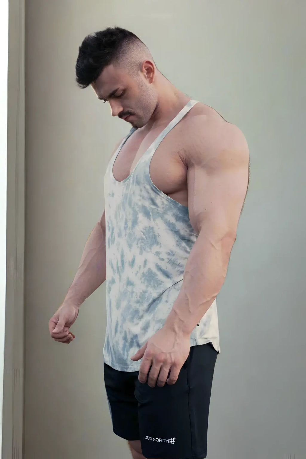 A man wearing a jed north classic bodybuilding racerback stringer tank top in gray tie dye with black shorts