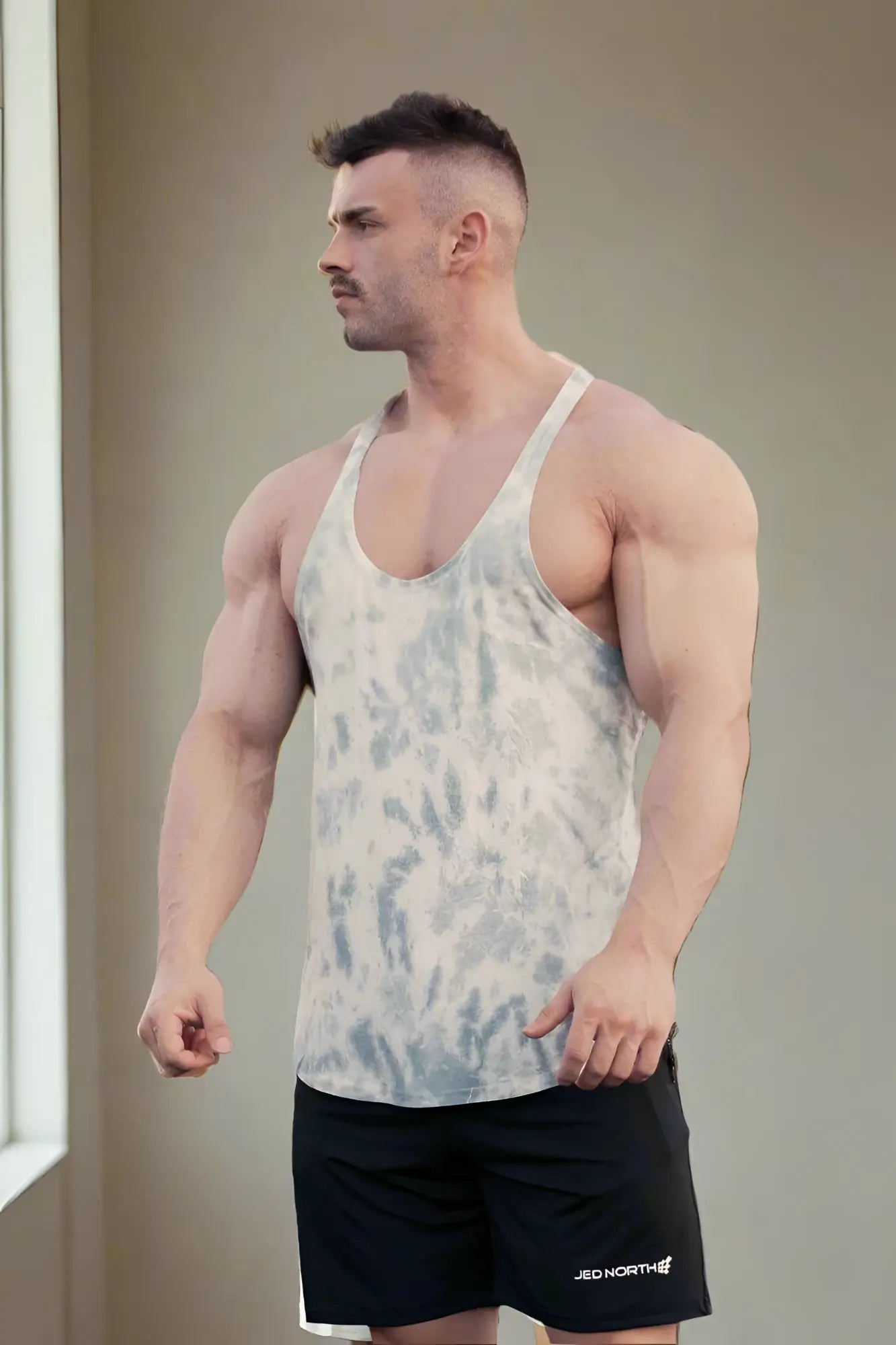 A muscular man wearing a gray tie dye Jed North classic bodybuilding racerback stringer with black shorts