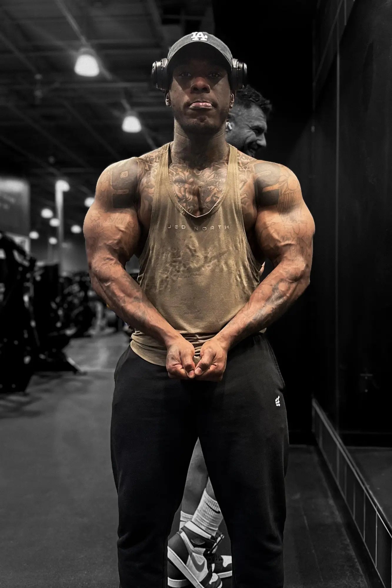 Muscular man wearing an olive Jed North classic bodybuilding racerback stringer tank top with black athletic pants and headphones