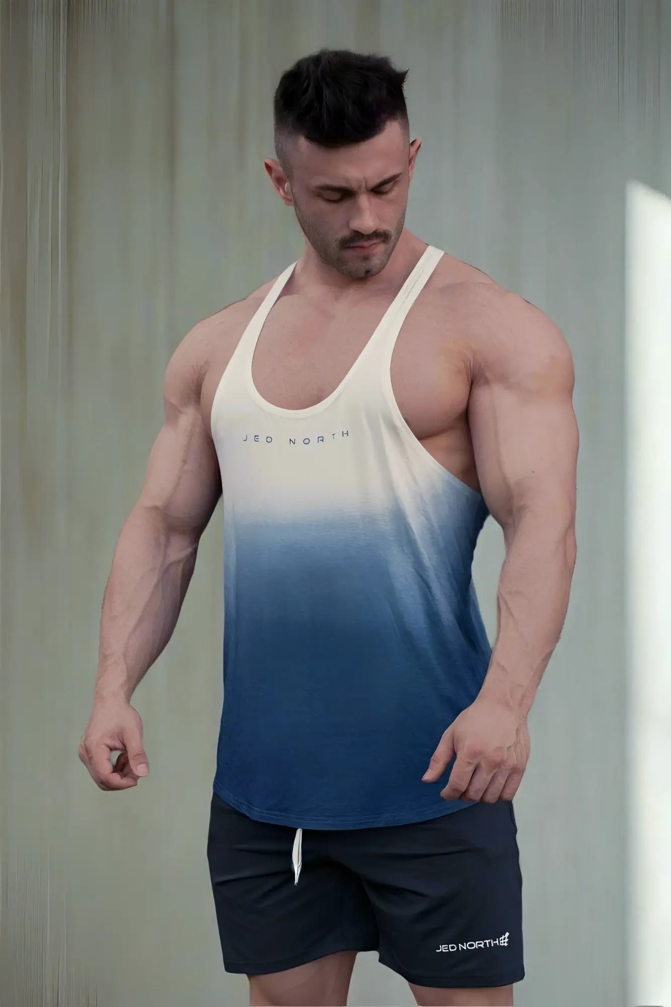 A men’s classic bodybuilding racerback stringer tank top in cream to navy blue ombre with subtle jed north logo and matching black shorts
