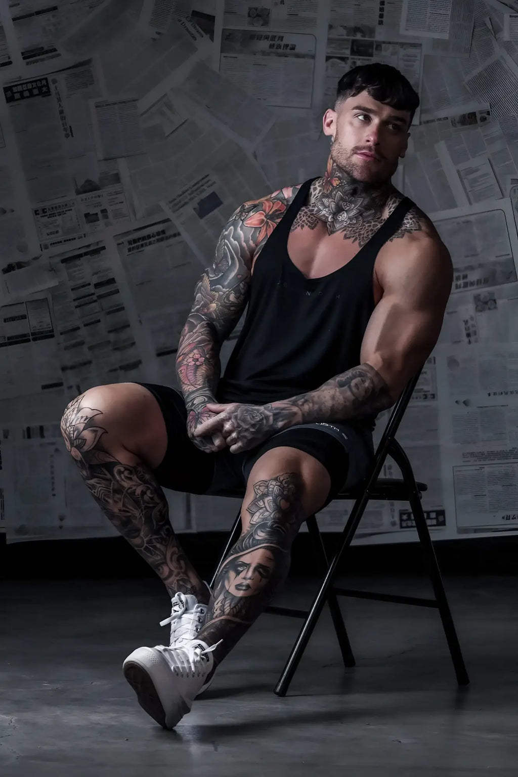 A muscular man with tattoos wearing a jed north classic bodybuilding racerback stringer in premium black sits on a chair