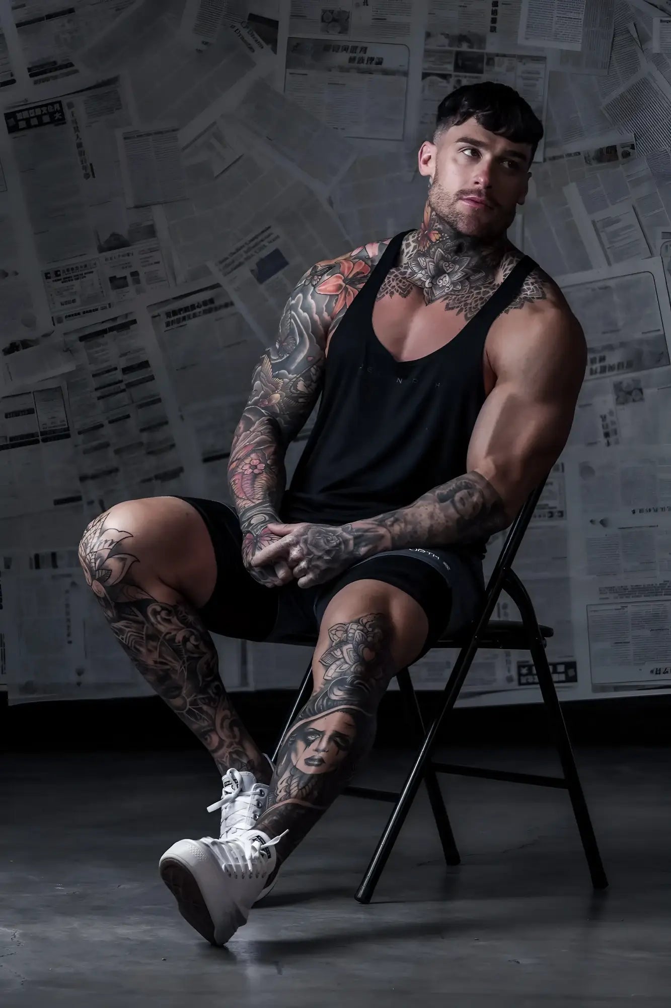 A muscular man with tattoos wearing a jed north classic bodybuilding racerback stringer in premium black sits on a chair