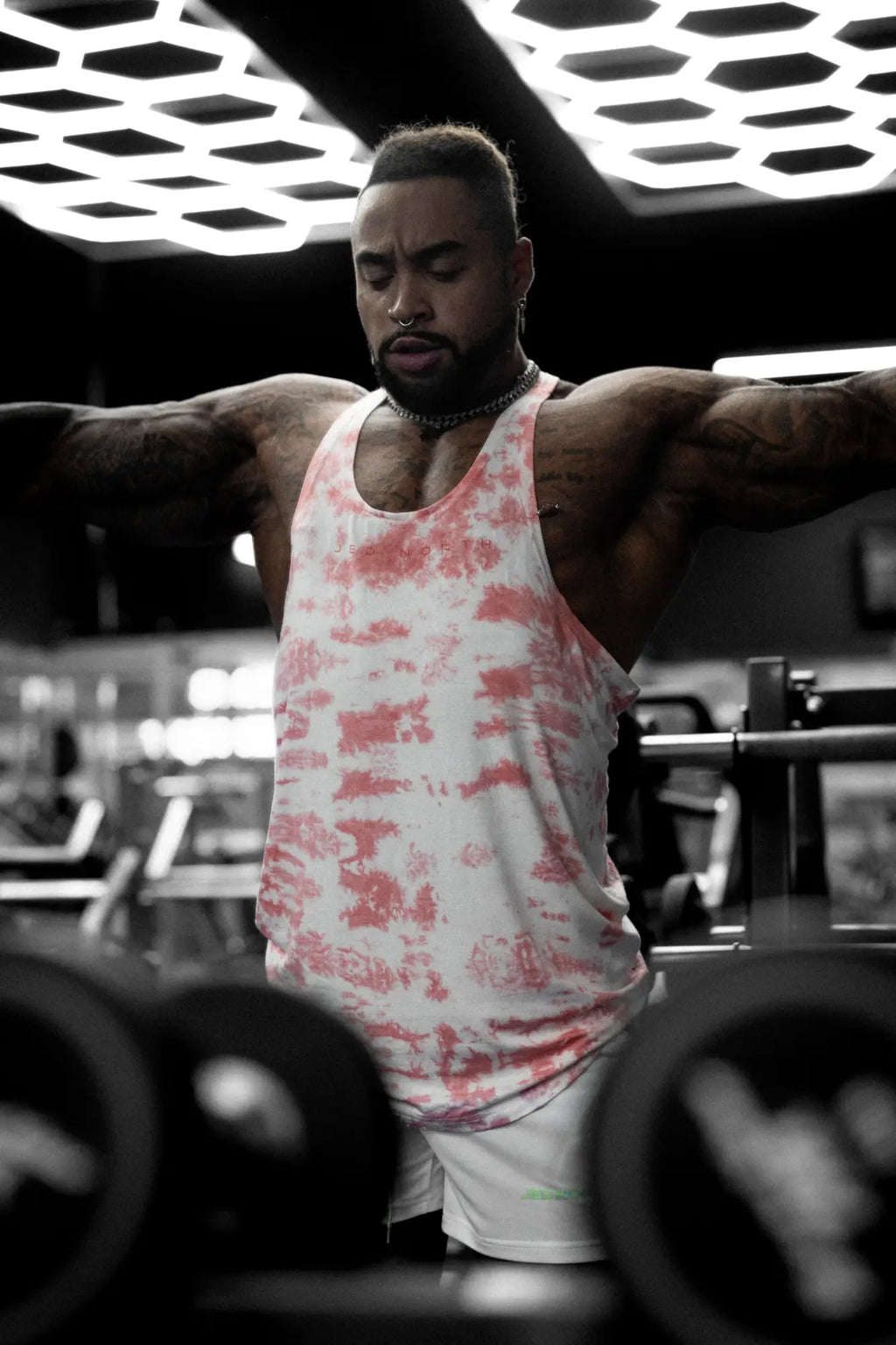 A man wearing a red tie dye classic bodybuilding racerback stringer tank top with visible arm tattoos