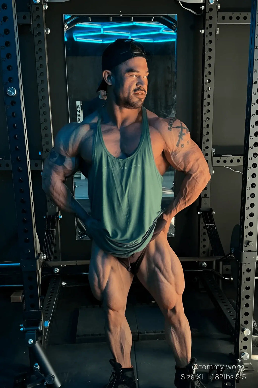 A muscular man wearing a sea green classic bodybuilding racerback stringer and black shorts in front of weightlifting rack
